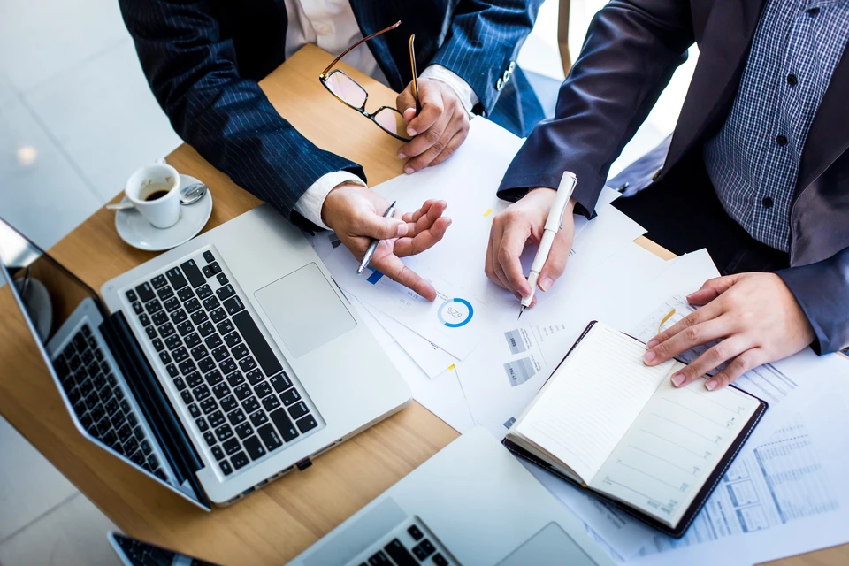 Réunion d'affaires en intérieur avec trois professionnels en costumes analysant des documents financiers sur table en bois